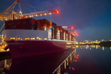 Pink container ship at night at Southampton Docks, UK. Aerial view of the bow and side view of the vessel loaded with shipping containers. Calm sea with ship and port crane lights reflection.